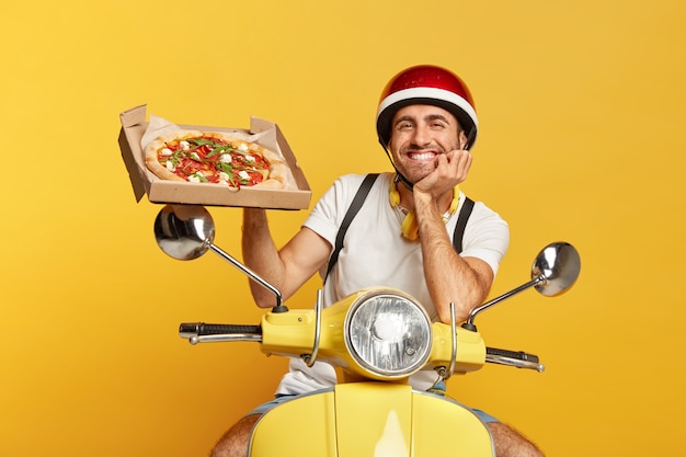 Free photo friendly looking deliveryman with helmet driving yellow scooter while holding pizza box