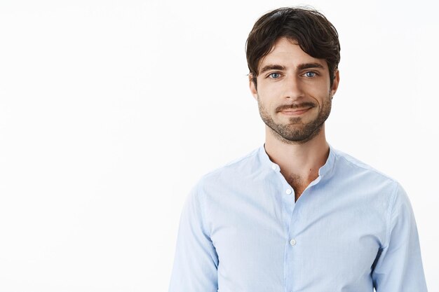 Friendly-looking optimstic and handsome mature and masculine male with blue eyes and bristle smiling joyfully as standing casually in blue shirt against gray wall