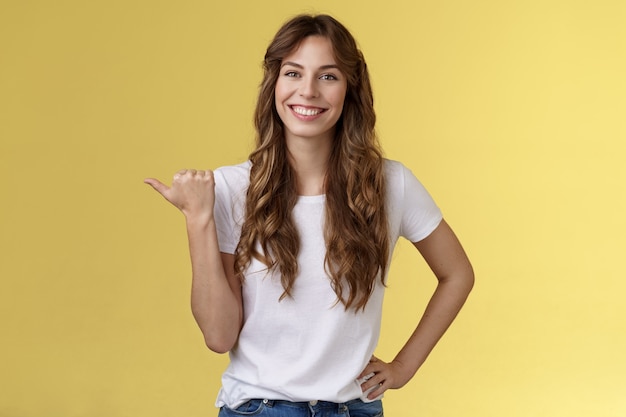 Friendly outgoing cheerful european cute female giving tips showing direction lively smiling toothy happily have nice pleasant conversation pointing left thumb introduce promo yellow background