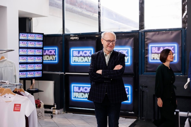 Free Photo friendly senior retail business owner wearing formal suit standing in clothing store against black friday neon banners, smiling man greeting and inviting customers to buy clothes during seasonal sales