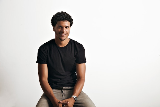 Friendly smiling young man in unlabeled cotton black t-shirt and jeans sitting on chair isolated on white