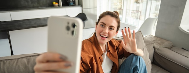 Friendly smiling young woman sitting on couch and waving hand at smartphone camera video chats