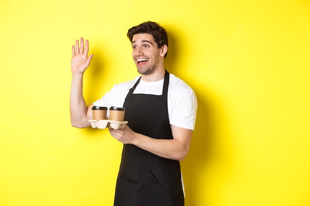 Friendly waiter in cafe waving hand at customer, holding takeaway coffee oder, standing against yellow background in black apron.