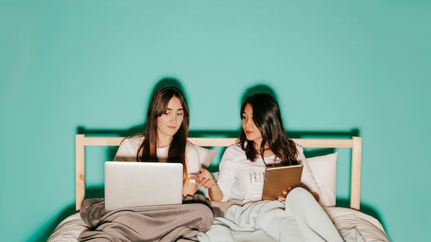 Friends browsing laptop and tablet in bed