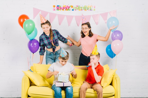 Free photo friends having fun while boy sitting on sofa unwrapping birthday gifts at home