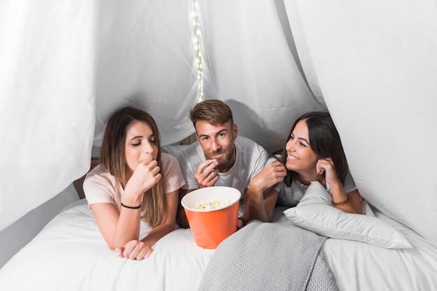 Friends lying on white cozy bed eating popcorns