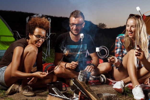 Friends sitting near bonfire, smiling, playing guitar Camping grill marshmallow.