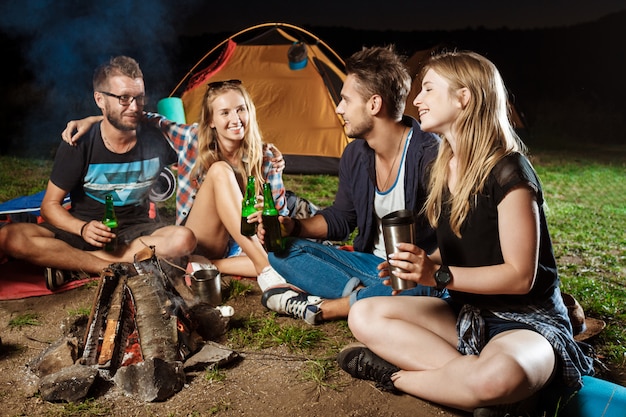 Friends sitting near bonfire, smiling, speaking, resting, drinking bear