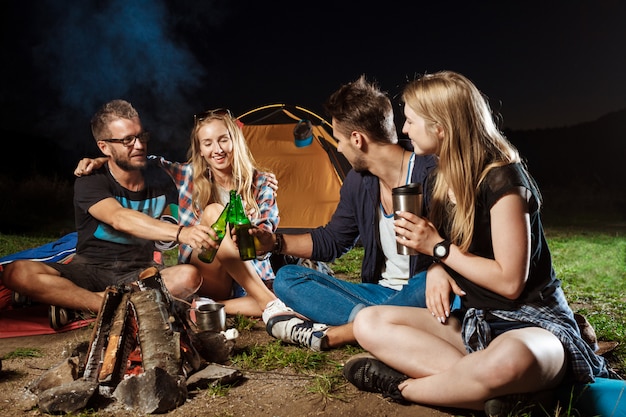 Friends sitting near bonfire, smiling, speaking, resting, drinking bear