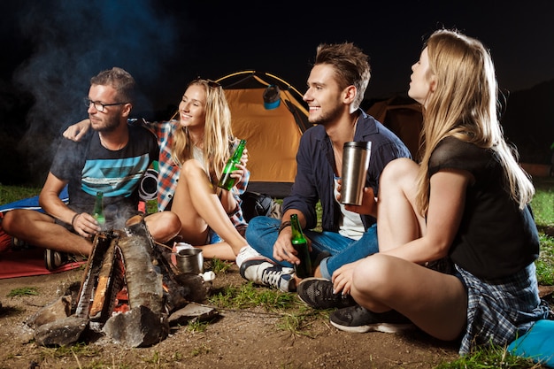 Friends sitting near bonfire, smiling, speaking, resting, drinking bear