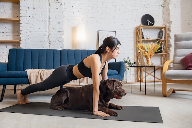 Friends. Young woman working out at home , doing yoga exercises with the dog. Beautiful woman stretching, practicing. Wellness, wellbeing, healthcare, mental health, lifestyle concept.