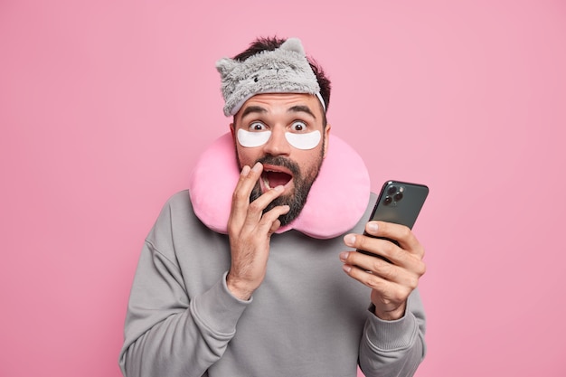 Frightened bearded adult man looks terrified, holds modern smartphone, wears sleepmask comfortable pillow around neck