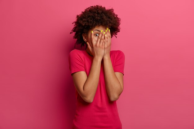 Frightened curly haired woman covers face with palms, afraids of something, expresses fear, stares and peeks through fingers, hides herself, wears casual t shirt, poses against rosy wall