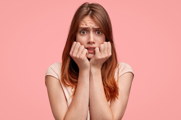 Frightened female with freckles on face, looks with terrified expression wears casual striped t shirt, isolated on pink background.