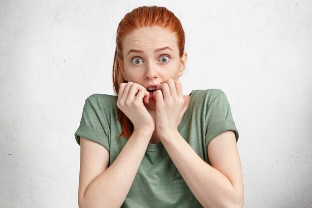 Frightened ginger woman with freckled skin stares at camera and bites nails, afraids of darkness, dressed in casual clothing, poses against white concrete wall.