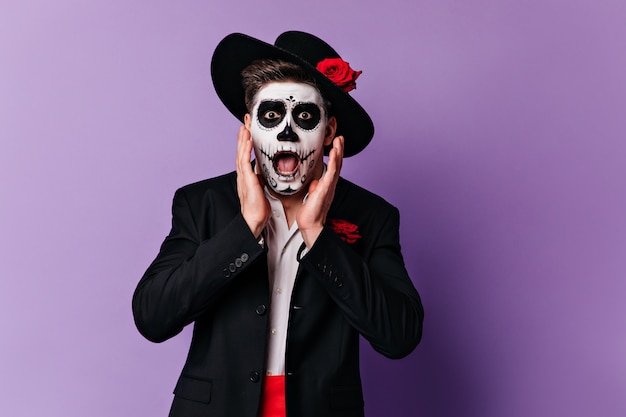 Frightened man in wide-brimmed hat looking in horror at camera. Portrait of guy with Halloween makeup posing on purple background.