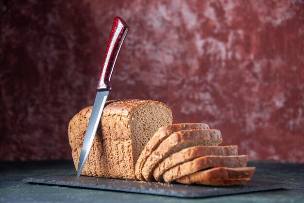 Front close view of black bread slices knife on dark color board on mixed colors distressed background