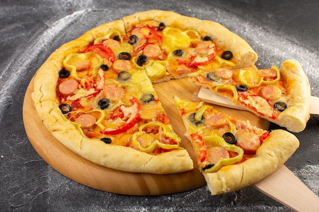 Front close view cheesy pizza with red tomatoes, black olives, bell-peppers and sausages on the dark desk