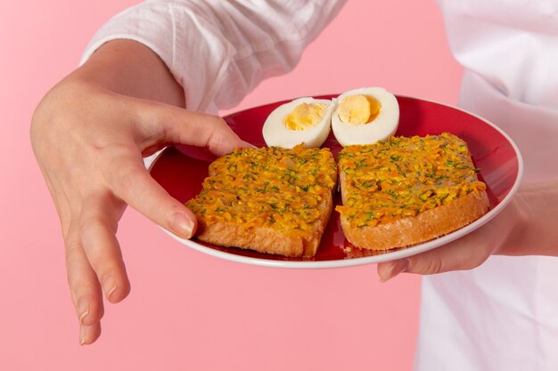 Front close view female confectioner in white wear holding plate with toasts and eggs on pink wall cook job kitchen cuisine food