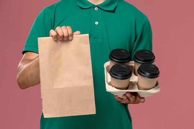 Front close view male courier in green uniform holding brown coffee cups and food package on pink desk