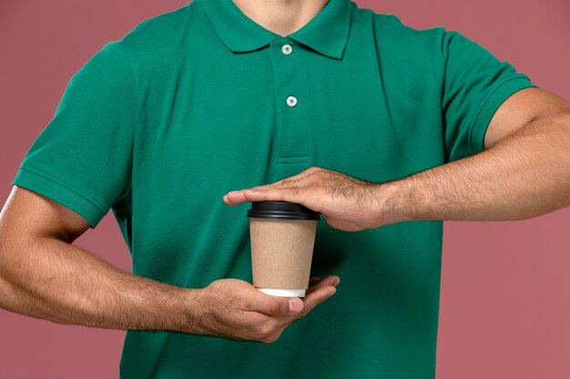 Front close view male courier in green uniform holding delivery coffee cup on the light-pink background background