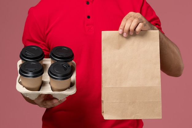 Front close view male courier in red uniform holding brown delivery coffee cups with food package on the pink wall service delivery job worker uniform