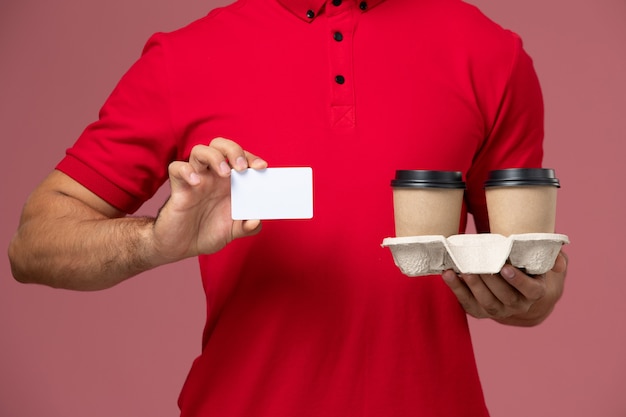 Free photo front close view male courier in red uniform holding brown delivery coffee cups with white card on pink wall