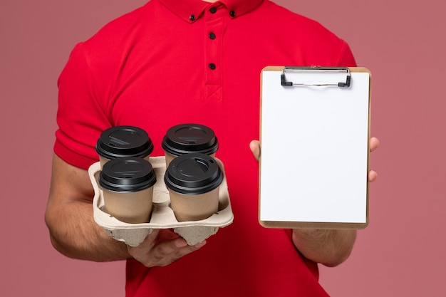 Free Photo front close view male courier in red uniform holding delivery coffee cups with notepad on the pink wall 