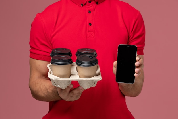 Free Photo front close view male courier in red uniform smiling and holding delivery coffee cups with phone on pink wall 