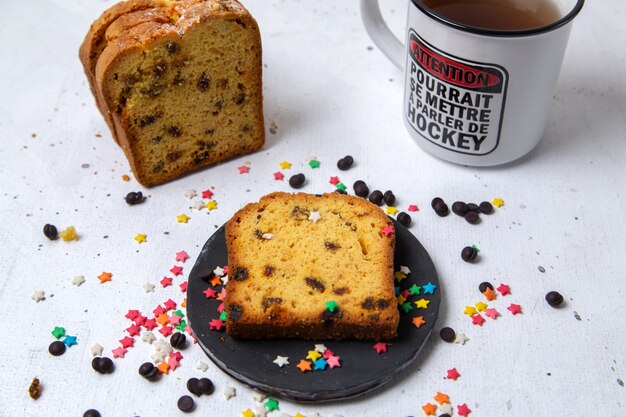 Front close view slice of cake inside dark plate with colorful candies and cup of tea