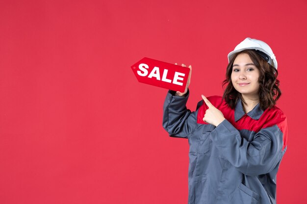 Free Photo front close view of smiling female worker in uniform wearing hard hat and pointing sale icon on isolated red wall