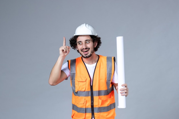 Front close view of smiling male construction worker in warning vest with safety helmet and showing blank pointing up on gray wall