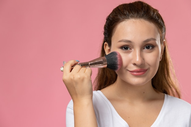 Front close view young attractive female in white t-shirt doing a make-up with a slight smile on the pink background