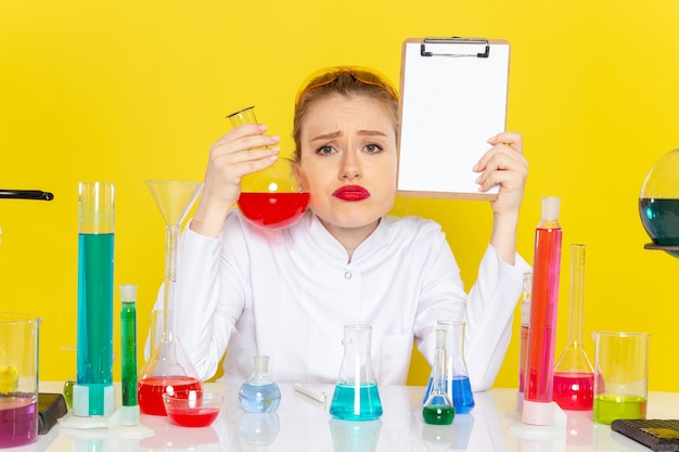 Front close view young female chemist in white suit with colored solutions working with them and sitting on yellow