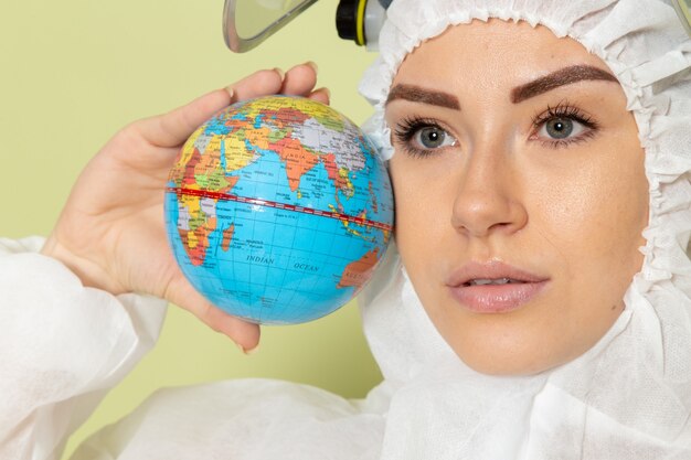 Front close view young female in white special suit and yellow helmet holding little globe on green