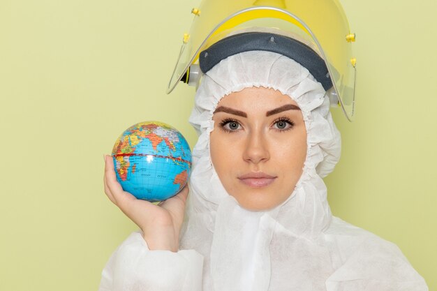 Front close view young female in white special suit and yellow helmet holding little globe on green