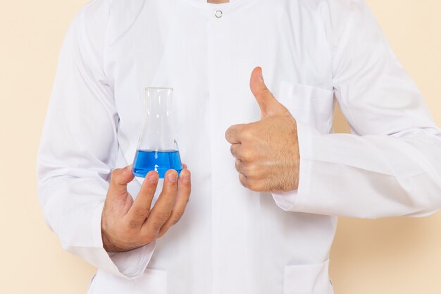 Front close view young male chemist in white special suit holding a little flask with blue solution on cream wall science experiment chemistry scientific