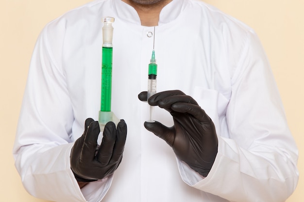 Free photo front close view young male chemist in white special suit holding little flask with green solution and injection on the cream wall science experiment lab