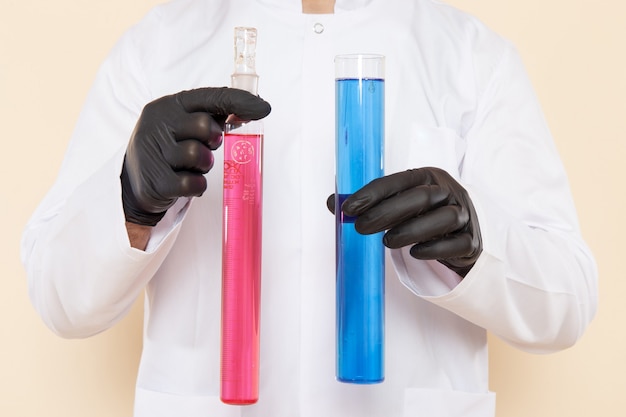 Front close view young male chemist in white special suit holding little flasks with colored solutions on cream wall lab chemistry science experiment