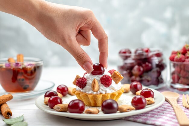 Front closer view little creamy cake with raspberries cherries and little biscuits taking by female on light desk