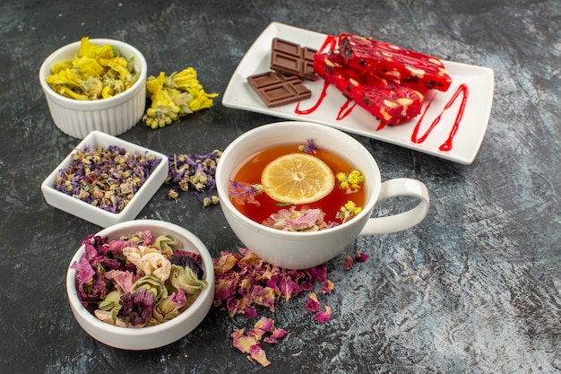 Free photo front shot of a cup of herbal tea with bowls of dry flowers and a plate of chocolate on grey ground