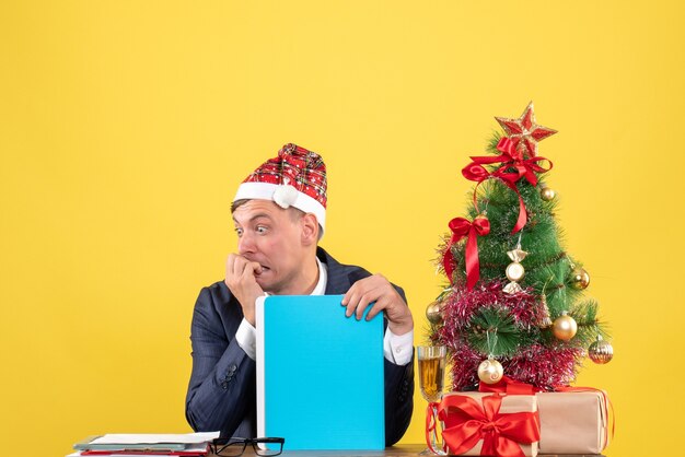 Front view of agitated business man sitting at the table near xmas tree and presents on yellow wall