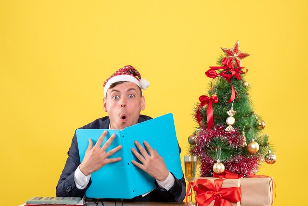 Front view of amazed business man sitting at the table near xmas tree and presents on yellow wall