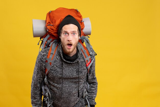 Front view of amazed male hitchhiker with leather gloves and backpack standing on yellow wall