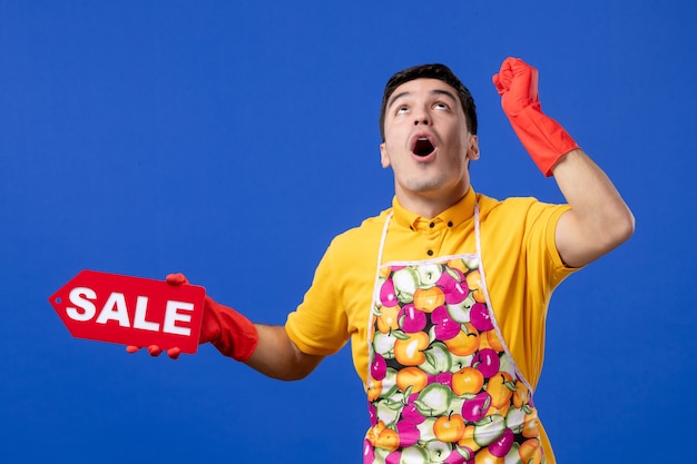 Front view amazed male housekeeper in yellow t-shirt holding sale sign looking up with surprise on blue space