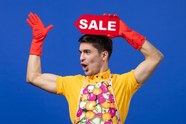 Front view amazed male housekeeper in yellow t-shirt raising sale sign over his head on blue space