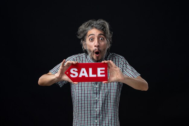 Front view amazed man holding up sale sign with both hands standing on dark wall