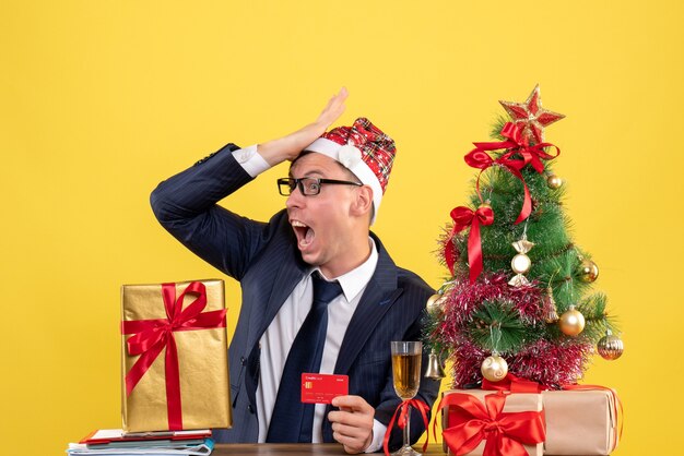 Front view amazed man putting hand to his forehead sitting at the table near xmas tree and presents on yellow background