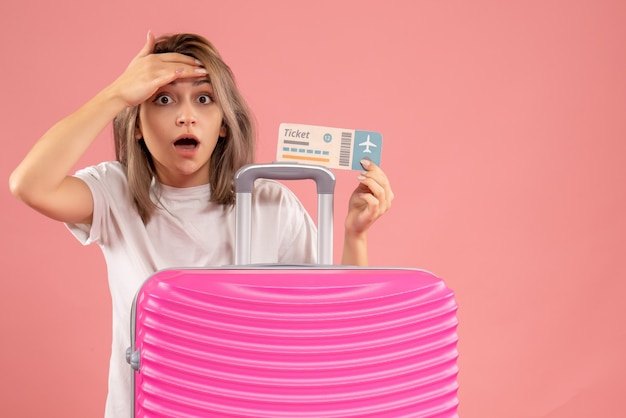 Front view of amazed young girl with pink suitcase holding ticket