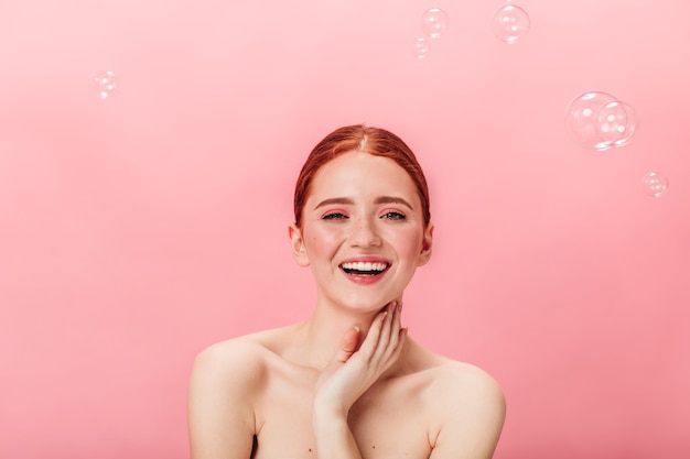 Front view of amazing woman with soap bubbles. Studio shot of happy nude ginger girl laughing on pink background.